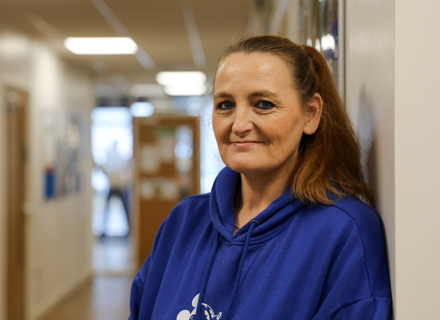 A middle-aged woman, wearing a blue hoodie, stands near a hallway wall, smiling gently towards the camera.