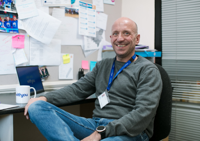 A smiling man sits at a desk in an office filled with papers and photographs, wearing a grey sweater and a blue lanyard.