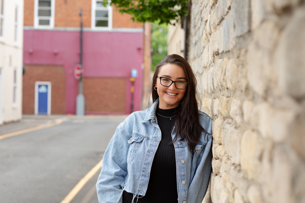 A cheerful young woman with glasses, wearing a denim jacket, stands by a stone wall on a street, with a red building in the background.