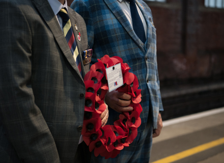 Two individuals in formal attire stand at a train station; one holds a wreath of red poppies and the other displays a medal and a ticket.