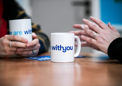 Two people sitting at a table, holding mugs with the text "WithYou" visible on one of them.