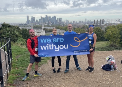 Four runners holding a "we are with you" banner with a city skyline in the background, standing atop a hill with a cloudy sky overhead.