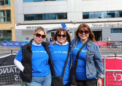 Three women wearing blue t-shirts and sunglasses stand together at an outdoor event, smiling at the camera.