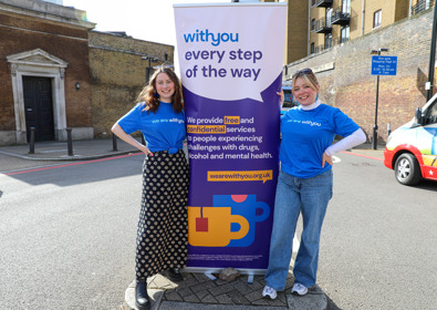 Two women in blue t-shirts stand smiling beside a banner that reads "with you every step of the way," promoting services for mental health and addiction.