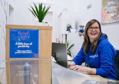A woman in a blue hoodie smiling at a reception desk with a laptop, motivational signs, and a plant in a modern office setting.