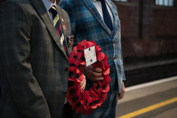 Two individuals in formal attire stand at a train station; one holds a wreath of red poppies and the other displays a medal and a ticket.